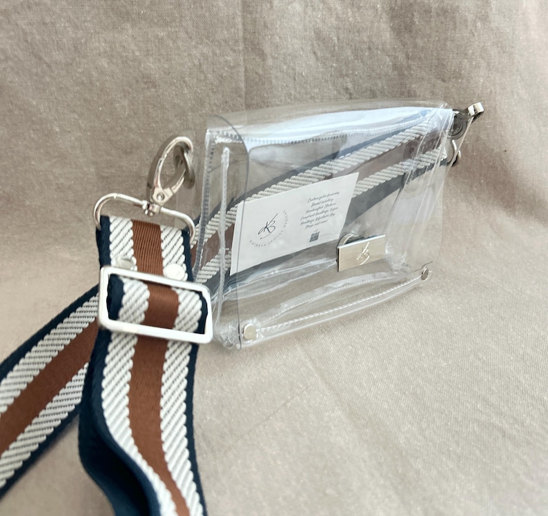 May include: A clear rectangular crossbody bag with silver-tone hardware and a detachable woven strap featuring navy blue, white, and brown stripes. A small white rectangular tag with black text and a logo is visible inside the transparent bag.