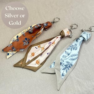 Könnte beinhalten: Drei Stoffschlüsselanhänger mit Metallringen, verziert mit Blumenmustern in Rost, Creme, Rosa und Hellblau. Jeder Schlüsselanhänger hat ein geknotetes, seidenartiges Banddesign. Der Text "Choose Silver or Gold" ist oben links sichtbar.