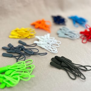 May include: A collection of colorful zipper pulls in various shades, including yellow, orange, blue, red, green, white, and gray. Each pull has a cord loop and a matching colored plastic or rubber grip. The pulls are arranged on a neutral-toned fabric.