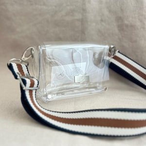 May include: A clear rectangular crossbody bag with a silver metal clasp closure. The bag features a wide, adjustable shoulder strap with horizontal stripes in navy blue, white, and brown. A small white label with text is visible inside the bag.