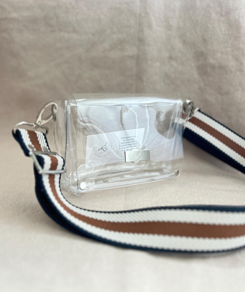 May include: A clear rectangular crossbody bag with a silver metal clasp closure. The bag features a wide, adjustable shoulder strap with horizontal stripes in navy blue, white, and brown. A small white label with text is visible inside the bag.