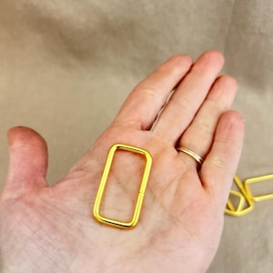 Puede incluir: Una hebilla rectangular de metal dorado sostenida en una mano. La hebilla tiene esquinas redondeadas y una abertura en un lado. Otra hebilla es parcialmente visible en el fondo. El fondo es de color marrón claro.