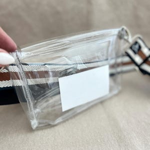 May include: A clear rectangular fanny pack featuring a top zipper closure and a white rectangular label on the front. The bag is attached to a wide, adjustable woven strap with horizontal stripes in shades of brown, white, and dark blue.