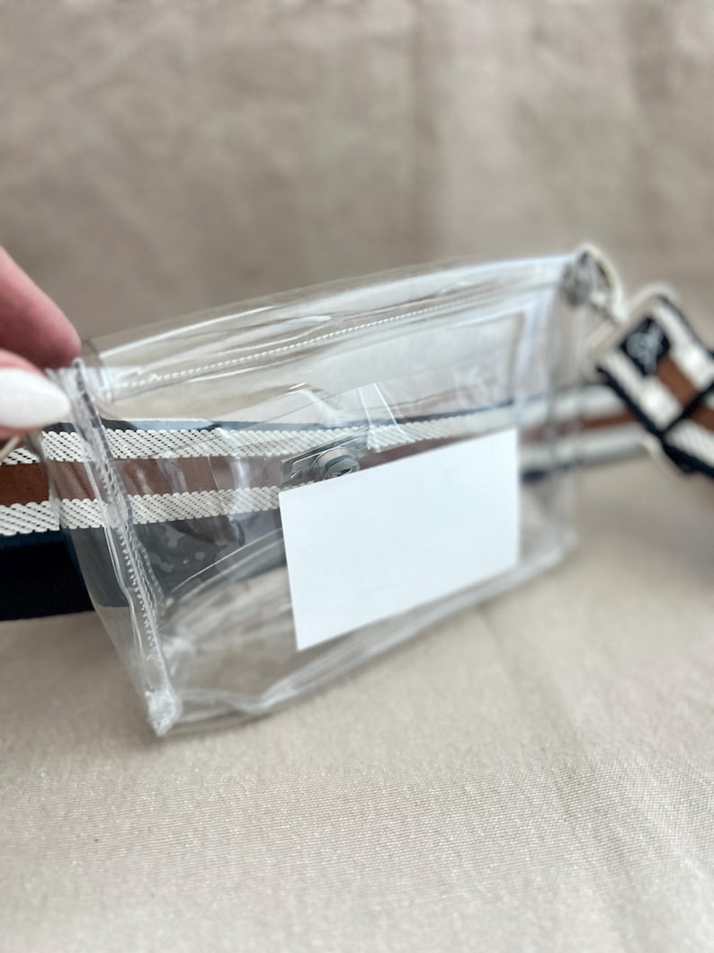 May include: A clear rectangular fanny pack featuring a top zipper closure and a white rectangular label on the front. The bag is attached to a wide, adjustable woven strap with horizontal stripes in shades of brown, white, and dark blue.