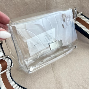 May include: A clear rectangular crossbody bag with a silver-toned metal turn-lock closure featuring a small bee emblem. The bag includes a wide, adjustable shoulder strap with horizontal stripes in navy blue, white, and brown.