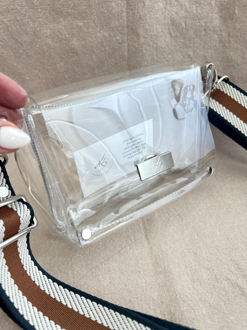 May include: A clear rectangular crossbody bag with a silver-toned metal turn-lock closure featuring a small bee emblem. The bag includes a wide, adjustable shoulder strap with horizontal stripes in navy blue, white, and brown.