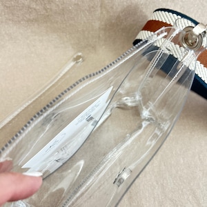 May include: A clear plastic crossbody bag with a white interior tag. The bag features a thick, woven shoulder strap with horizontal stripes in dark blue, white, and brown. The bag has a rectangular shape with rounded corners and silver hardware.