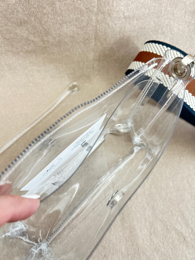 May include: A clear plastic crossbody bag with a white interior tag. The bag features a thick, woven shoulder strap with horizontal stripes in dark blue, white, and brown. The bag has a rectangular shape with rounded corners and silver hardware.