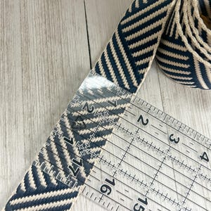 May include: A navy blue and cream woven fabric strap featuring a diagonal chevron pattern. A clear plastic ruler is placed over the strap, showing a width of approximately 1.5 inches. The strap is coiled at one end and includes a metal hardware piece.