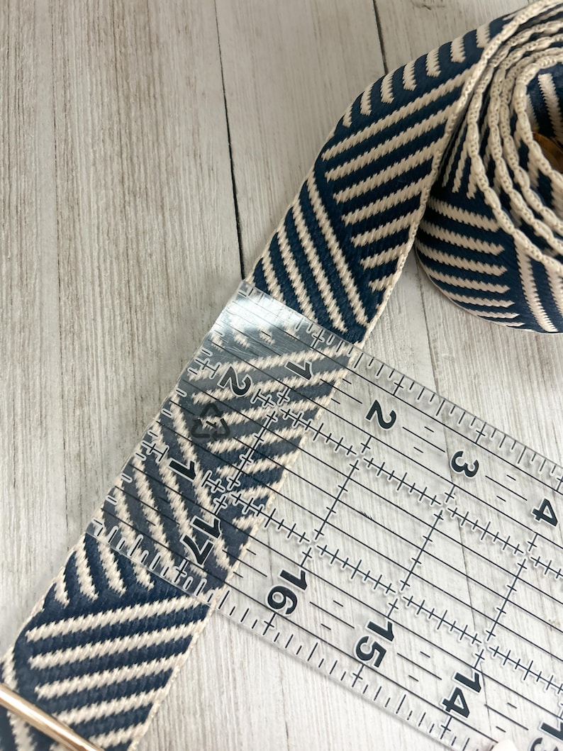 May include: A navy blue and cream woven fabric strap featuring a diagonal chevron pattern. A clear plastic ruler is placed over the strap, showing a width of approximately 1.5 inches. The strap is coiled at one end and includes a metal hardware piece.
