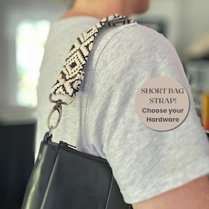 Könnte beinhalten: Ein kurzer Taschengurt mit einem cremefarbenen und schwarzen geometrischen Webmuster, befestigt an einer schwarzen Tasche mit einem Metallkarabiner. Eine kreisförmige Grafik zeigt den Text: SHORT BAG STRAP! Choose your Hardware.