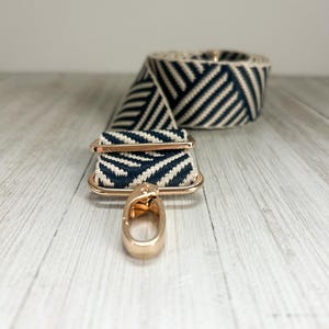 May include: A replacement bag strap featuring a navy blue and cream chevron pattern woven fabric. The strap includes polished gold-tone metal hardware, including a rectangular sliding adjuster and a swivel lobster clasp hook at the end.
