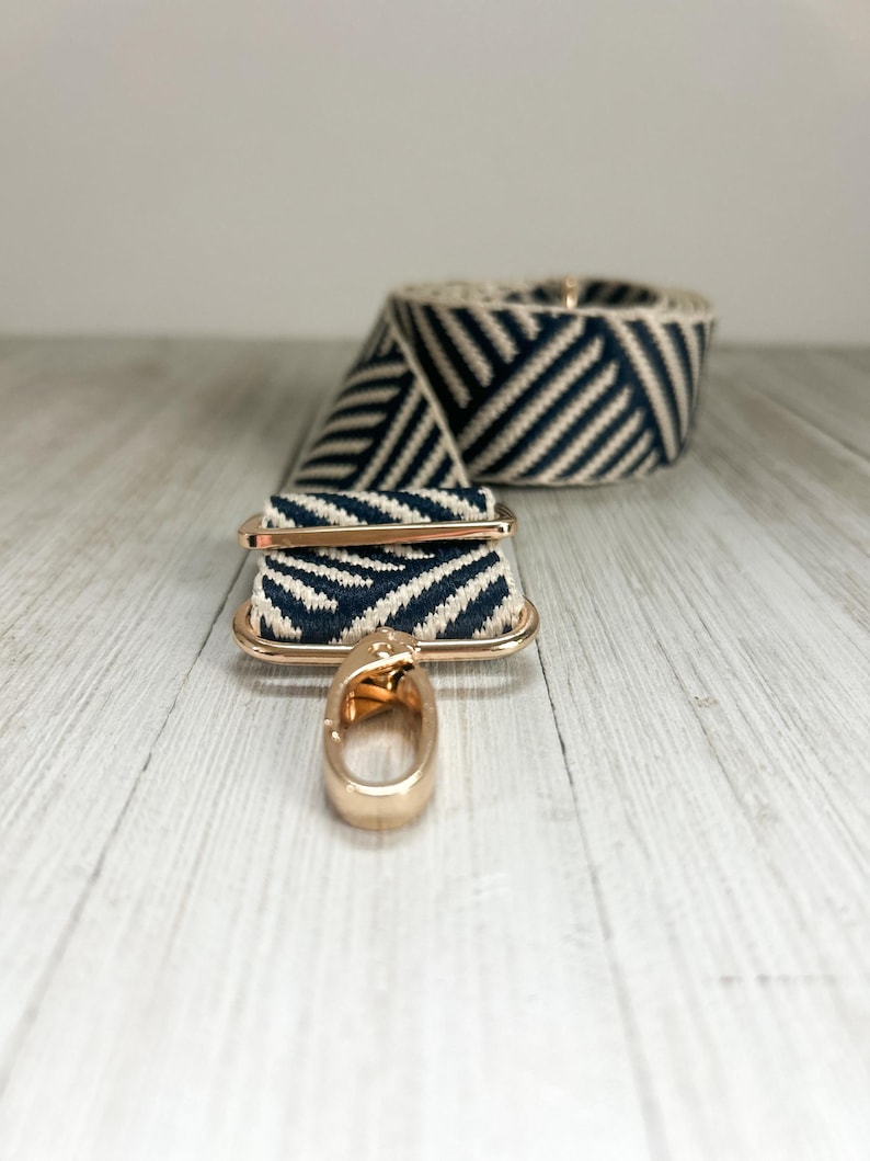 May include: A replacement bag strap featuring a navy blue and cream chevron pattern woven fabric. The strap includes polished gold-tone metal hardware, including a rectangular sliding adjuster and a swivel lobster clasp hook at the end.