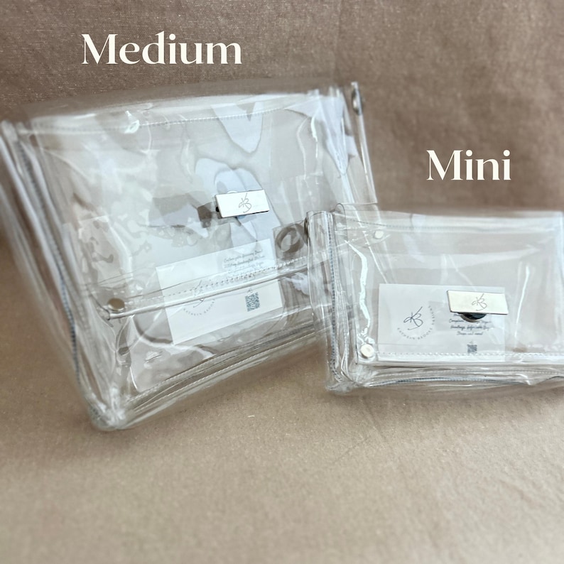 May include: Two clear rectangular pouches labeled Medium and Mini. Each pouch features a silver-toned metal clasp closure on the front flap and a small white rectangular tag with black text and a logo visible inside the transparent material.
