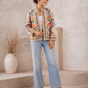Peut inclure: Veste rose clair brodée avec un col mandarin et un motif floral. La veste présente des broderies orange, bleues et vertes. Le modèle porte un jean bleu clair et des talons blancs.