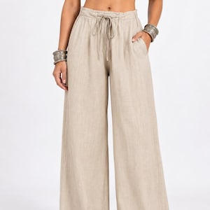 Puede incluir: Pantalones de color beige con pernera ancha, cintura alta, cinturilla elástica y cordón ajustable a juego. Estos pantalones largos incluyen bolsillos laterales y un corte holgado y relajado en las piernas, confeccionados en tela texturizada.