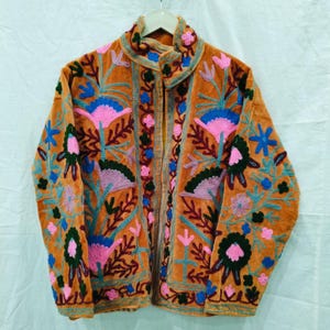May include: An orange velvet jacket with intricate floral embroidery in pink, blue, green, and purple. The jacket features a high collar and a button-up front, showcasing a bohemian style.