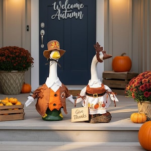 Op de afbeelding: Twee ganzenbeeldjes in herfstoutfits staan op een veranda. Eén gans draagt een bruine hoed en vest, de andere een witte jurk met een pompoenenschort. Een bordje leest "Give Thanks". Pompoenen en chrysanten op de achtergrond.