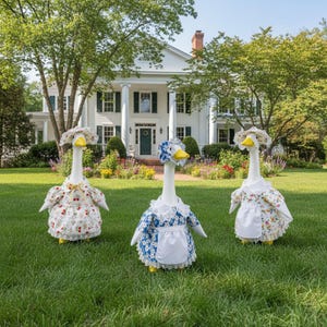 May include: Three goose ornaments, each wearing a floral dress, bonnet, and apron, are positioned on a green lawn before a white house with columns. The geese have yellow beaks and feet. The dresses display red, blue, and yellow floral designs.