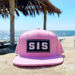 May include: A pink baseball cap with a black and white patch that says "SIS" on it. The cap is sitting on a sandy beach with a blurred background of the ocean and a palm tree.