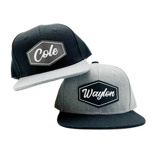 May include: Two flat-brimmed baseball caps. One is black with a gray brim and a white-lettered "Cole" patch. The other is gray with a black brim and a white-lettered "Waylon" patch. Both patches are in a black hexagon.