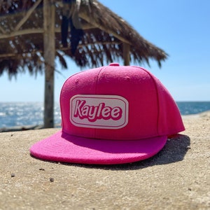 May include: A pink and white baseball cap with the word "Kaylee" in white letters on a white oval patch. The cap is sitting on a sandy beach.