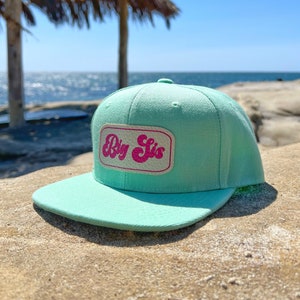 May include: A mint green flat-brimmed baseball cap with a rectangular patch that reads "Big Sis" in pink, retro-style lettering. The hat is set against a beach backdrop with a blue sky and ocean.