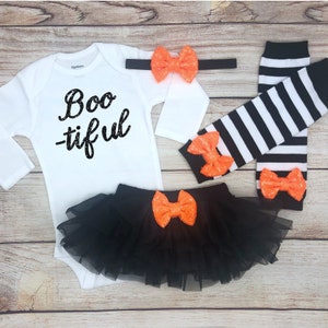 May include: A white long-sleeve bodysuit with the black text Boo-tiful, a black tulle skirt, a black headband, and two black and white striped leg warmers. Each piece is accented with a bright orange sequined bow for a coordinated set.