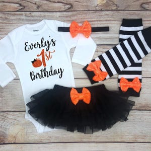 May include: A white long-sleeved baby bodysuit with the text "Everly's 1st Birthday" in black and orange. The set includes a black tutu, striped leg warmers, and a headband, all with orange bows.