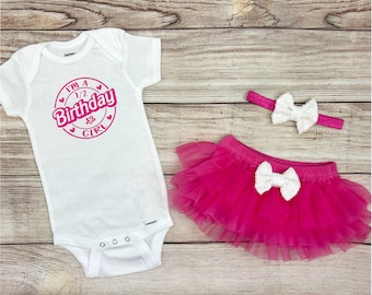 May include: A white baby bodysuit with the text "I'M A 1/2 Birthday GIRL" in pink, a hot pink tutu skirt, and a matching headband with a white bow. The outfit is designed for a baby's half-birthday celebration.