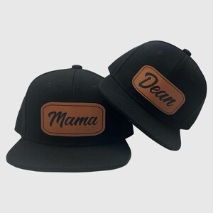 May include: Two black baseball caps with brown leather patches. The patch on the left cap says "Mama" and the patch on the right cap says "Dean".