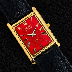 Könnte beinhalten: Eine rechteckige, goldfarbene Seiko Quartz Armbanduhr mit einem leuchtend roten Zifferblatt mit dezenter Wellenstruktur, goldfarbenen Zeigern und schwarzen römischen Ziffern. Die Uhr hat ein schwarzes, strukturiertes Lederarmband.