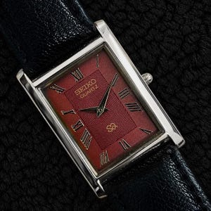 Op de afbeelding: Een rechthoekig Seiko Quartz polshorloge met een dieprode wijzerplaat met een golvend textuurpatroon, zilverkleurige Romeinse cijfers als uurmarkeringen en dunne zilveren wijzers. De horlogekast is van gepolijst zilverkleurig metaal met een zwarte leren band.