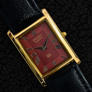 Peut inclure: Une montre-bracelet rectangulaire Seiko Quartz avec un boîtier doré, un cadran rouge profond à motif ondulé texturé et des chiffres romains noirs. La montre possède des aiguilles dorées et un bracelet en cuir noir à la texture grainée.