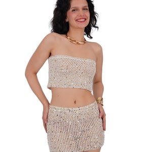 May include: A two-piece crochet set in a neutral color, consisting of a bandeau top and a mini skirt. The set is embellished with sequins. A gold-tone chain necklace and a gold bracelet are also visible.
