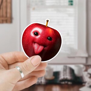 May include: A red apple sticker with a cartoon face, featuring black eyes and a pink tongue sticking out. The apple has a stem and is held by a hand with a silver ring.