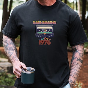 May include: Black t-shirt featuring a retro cassette tape design with the words "Rare Release" above and "1976" below. The design incorporates shades of brown, orange, and blue. The person is holding a dark blue mug.