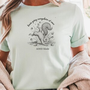 Op de afbeelding: Lichtgroen t-shirt met een zwart-wit illustratie van een vallende eekhoorn met eikels. De tekst luidt "brain going everywhere at once" en daaronder "ADHD Mode".