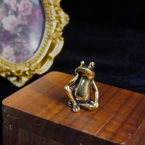 Puede incluir: Una pequeña figura de rana dorada en posición sentada, sobre una caja de madera. La rana tiene detalles visibles en ojos y extremidades. En el fondo, un espejo ovalado con marco dorado refleja un arreglo floral.