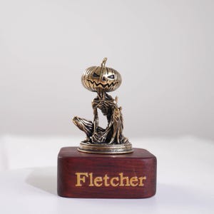 May include: A small bronze-toned figurine featuring a creature with a carved pumpkin head and a slender, textured body. The figure is mounted on a rectangular dark wood base with the name Fletcher engraved in gold lettering on the front face.