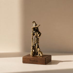 Puede incluir: Una figura de bruja de color bronce que sostiene una escoba y un libro, con un gato a sus pies. La bruja lleva un sombrero puntiagudo y un vestido largo. La figura está montada sobre una base de madera oscura con el nombre "Sophie" grabado.