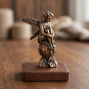 Könnte beinhalten: Bronze-farbene Feenfigur, sitzend auf einer Muschel, montiert auf einem Holzsockel. Die Fee hat detaillierte Flügel und befindet sich in einer nachdenklichen Pose. Auf dem Sockel steht das Wort "Fairy".