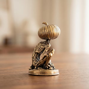 May include: A small, metallic, bronze-colored figurine depicting a seated, humanoid creature with a pumpkin-shaped head. The figure features intricate, textured, root-like details across its body and sits upon a circular, flat base.