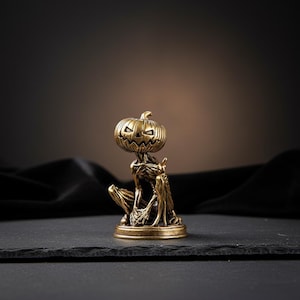 May include: A small, metallic gold-colored figurine depicting a skeletal creature with a carved pumpkin head. The figure is in a seated position, featuring intricate details on the ribcage, limbs, and the jack-o'-lantern face with triangular eyes and a jagged mouth, all set on a circular base.