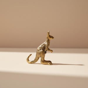 May include: A small, detailed brass kangaroo figurine. The kangaroo is standing upright with its tail curled and its arms at its sides. The figurine has textured fur and is set against a neutral background.