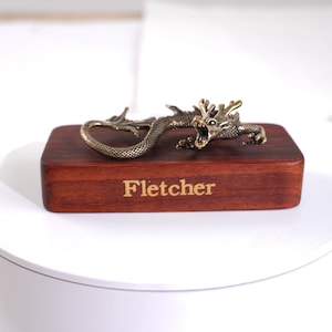 Op de afbeelding: Een metalen, messingkleurig drakenbeeldje met gedetailleerde schubben en een open bek, gemonteerd op een rechthoekig, donkerbruin houten blok. Op de voorzijde van het houten blok staat de naam Fletcher gegraveerd in een lichtgekleurd, schreefloos lettertype.