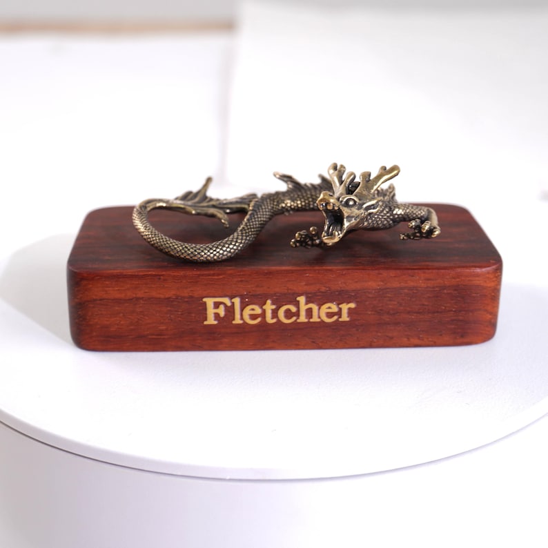 Op de afbeelding: Een metalen, messingkleurig drakenbeeldje met gedetailleerde schubben en een open bek, gemonteerd op een rechthoekig, donkerbruin houten blok. Op de voorzijde van het houten blok staat de naam Fletcher gegraveerd in een lichtgekleurd, schreefloos lettertype.