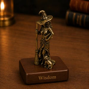 Puede incluir: Una figura de metal en tono bronce de una bruja con sombrero puntiagudo, sosteniendo una escoba y de pie junto a un gato. La figura está montada sobre una base de madera rectangular con la palabra Wisdom grabada en letras doradas.