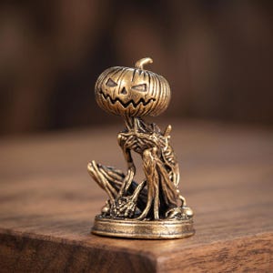 May include: A small brass-colored figurine depicting a creature with a carved pumpkin head featuring a jagged, smiling face. The body is slender and textured with root-like details, seated on a circular base with a metallic, antique finish.