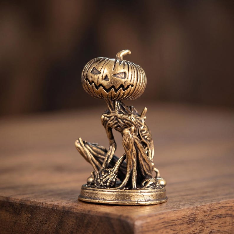 May include: A small brass-colored figurine depicting a creature with a carved pumpkin head featuring a jagged, smiling face. The body is slender and textured with root-like details, seated on a circular base with a metallic, antique finish.